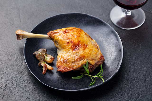 Pies de pato asado en un plato negro junto al vino.